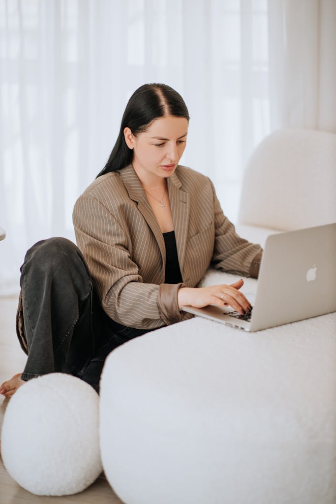 A young woman in business attire works on her laptop from a cozy indoor setting.